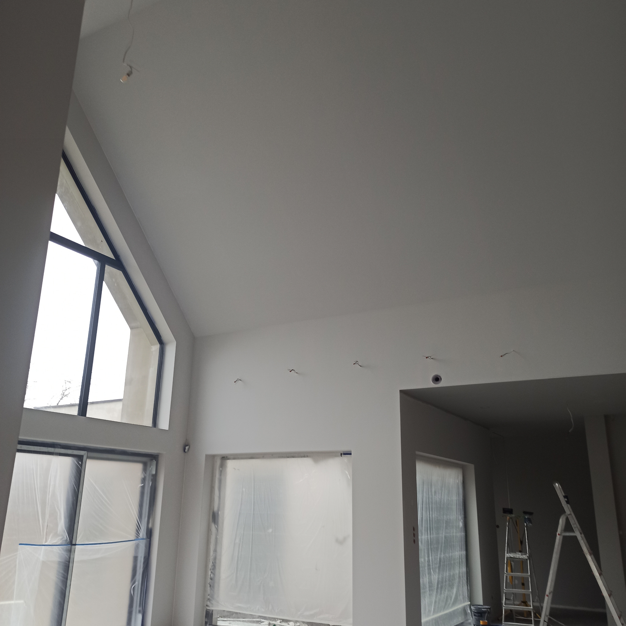 Wnętrze po świeżym pomalowaniu na biało, okna zabezpieczone folią malarską, widoczne kable wystające z sufitu i drabina malarska.