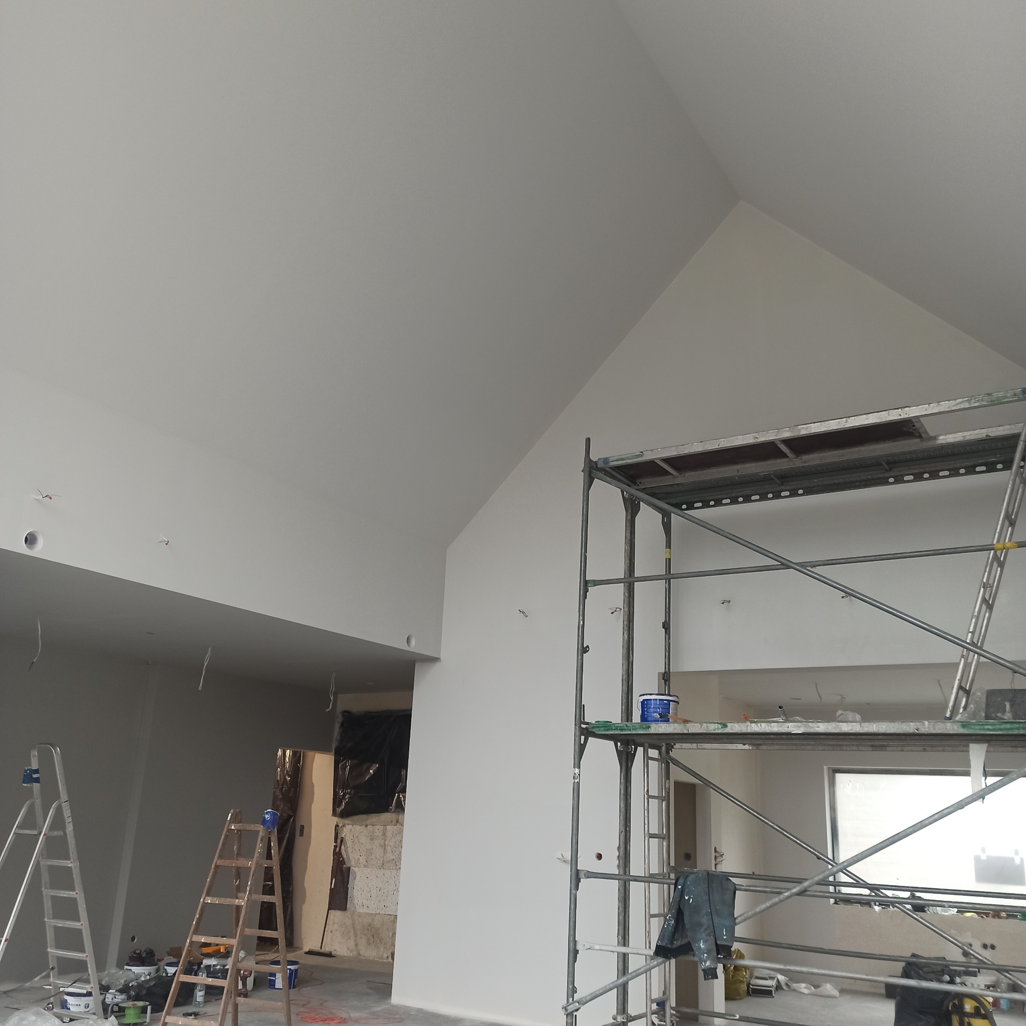 Wnętrze w trakcie prac wykończeniowych: świeżo pomalowane na biało ściany i sufit, widoczne rusztowanie z farbą, drabiny malarskie i zabezpieczone folią fragmenty ścian.