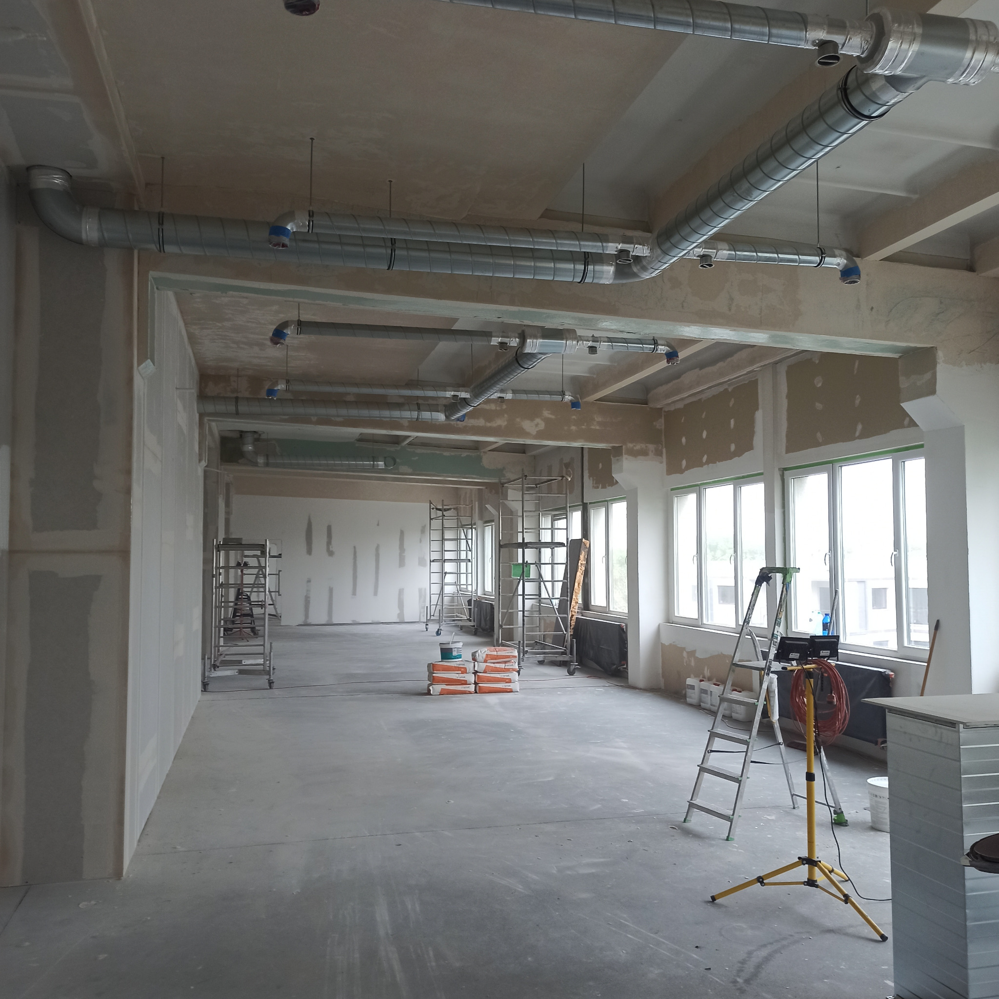 Przestronne wnętrze biurowe w trakcie prac wykończeniowych: ściany przygotowane pod nakładanie gładzi, widoczne rury wentylacyjne pod sufitem, drabiny i worki z materiałami budowlanymi.