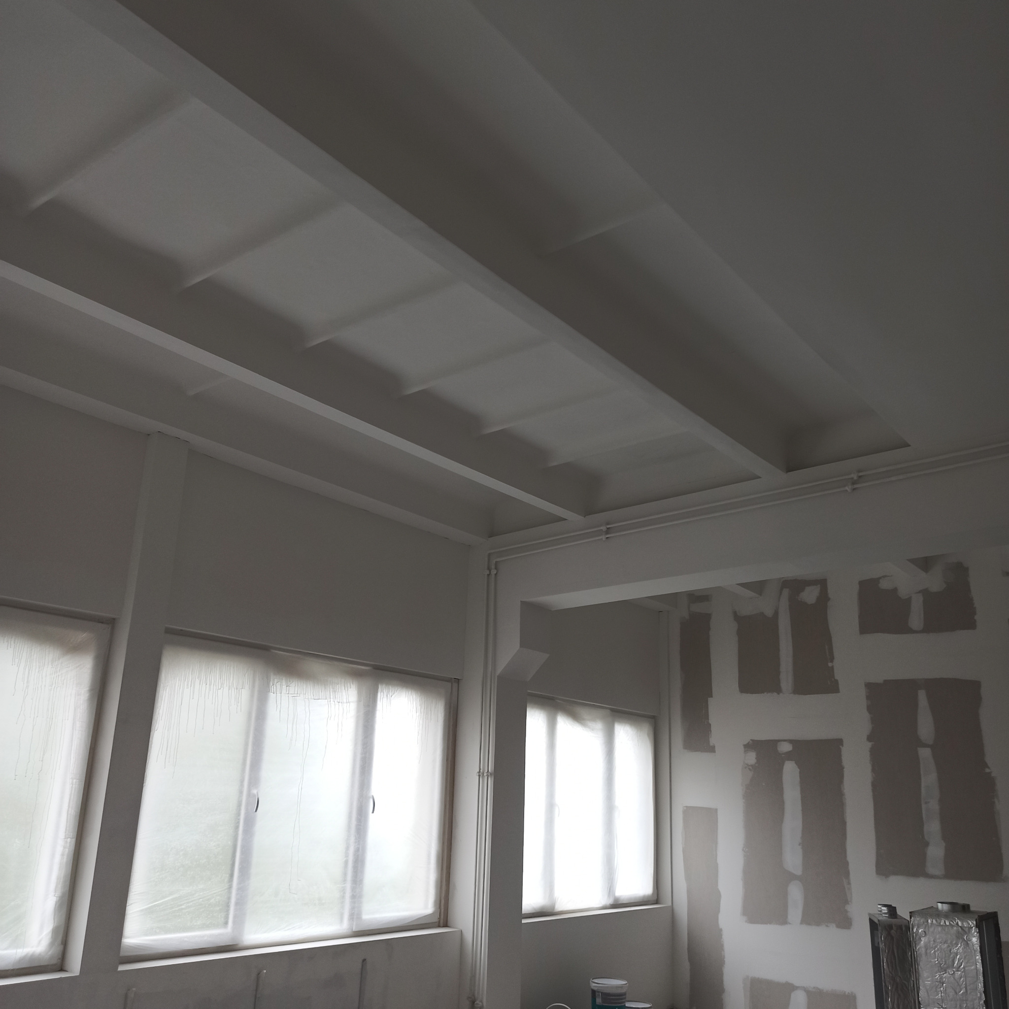 Wnętrze w trakcie prac przygotowawczych do malowania: okna zabezpieczone folią, ściany z widocznymi łatami gładzi, sufit z kasetonami pomalowany na biało, w rogu widoczne puszki z farbą i metalowe...