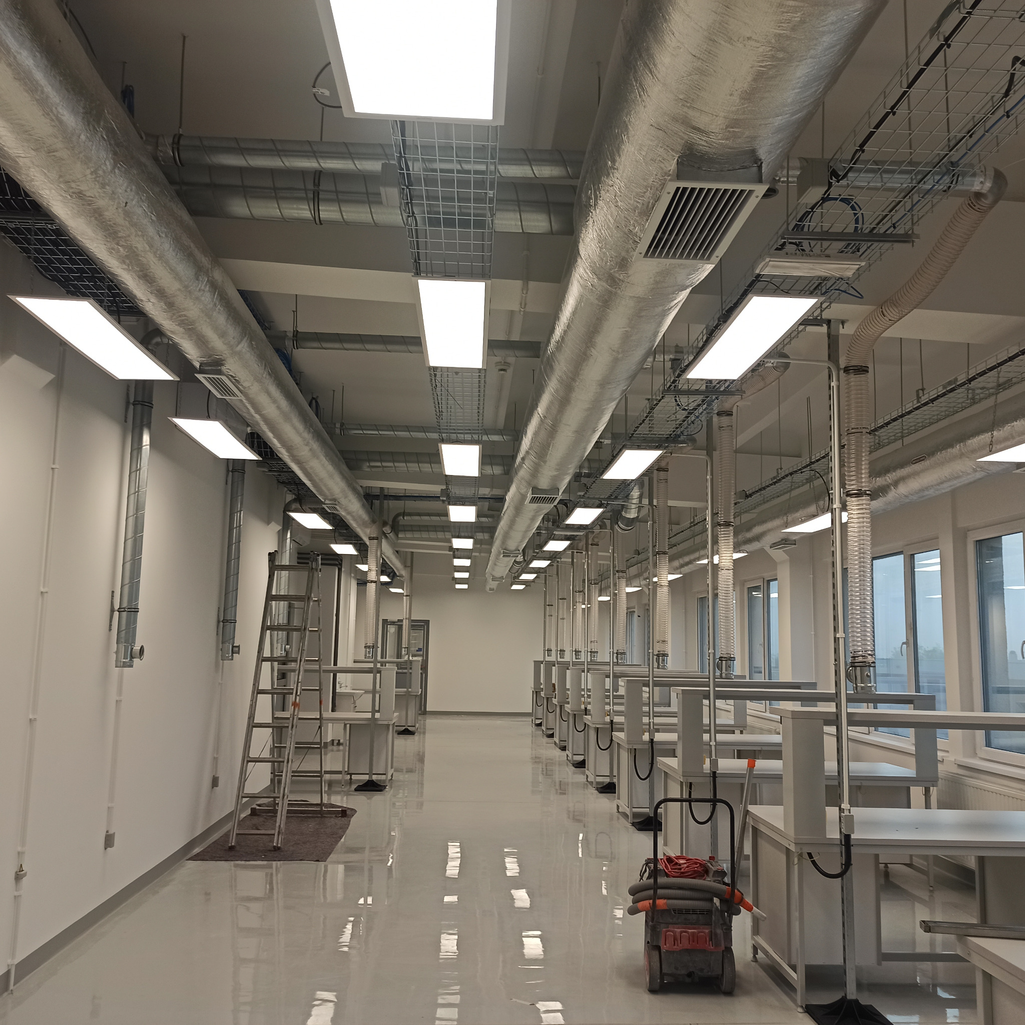 Wnętrze biura z instalacją wentylacyjną pod sufitem, widoczne metalowe rury, kratownice i drabina oparta o ścianę, podłoga pokryta żywicą epoksydową.