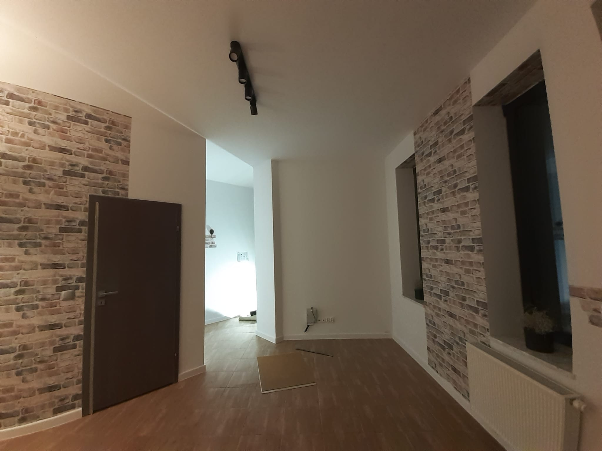 Pusty pokój po remoncie z panelami podłogowymi, ścianą z tapetą imitującą cegłę i białymi ścianami, widoczne gniazdka elektryczne i lampa sufitowa.