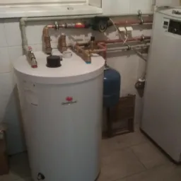 Bojler marki Saunier Duval z podłączonymi miedzianymi rurami i zaworami, widoczny również mały zbiornik wyrównawczy i pompa obiegowa, tło stanowi biała ściana z kafelkami.