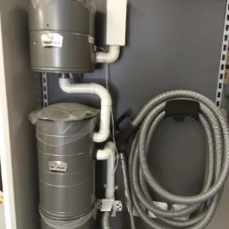 Szary, trzypoziomowy system odkurzacza centralnego VacuMaid z rurami PCV i zwiniętym wężem, zaprezentowany na tle szarej ściany z napisem 'ODKURZACZE CENTRALNE'.