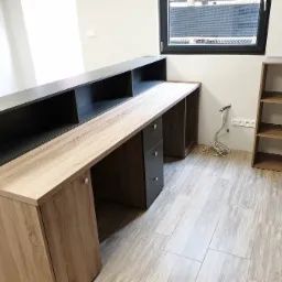 Biuro z blatem drewnianym, szafkami z płyty wiórowej imitującej drewno oraz czarnymi szufladami, widoczne okablowanie elektryczne i półka w tle.