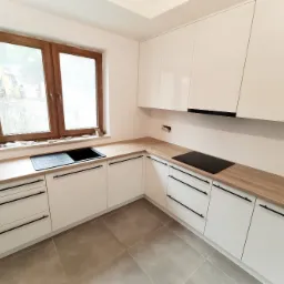 Nowoczesna, jasna kuchnia z białymi, lakierowanymi frontami szafek i drewnianym blatem. Widoczne czarne, minimalistyczne uchwyty, czarny zlew i płyta indukcyjna. Za oknem widoczny sprzęt budowlany.