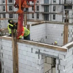 Dwóch robotników w odblaskowych kamizelkach obsługuje pomarańczowy lej do wylewania betonu, podczas budowy ścian z białych bloczków na placu budowy wielopiętrowego budynku.