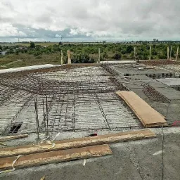 Zbrojenie stropu na budowie domu, widoczne pręty stalowe ułożone w siatkę, drewniane deski szalunkowe i słupy podtrzymujące konstrukcję, w tle krajobraz z zielenią i pochmurne niebo.