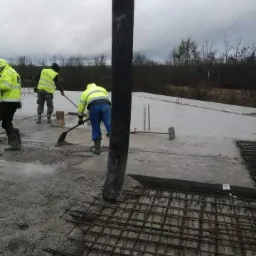 Wylewanie betonu na zbrojenie fundamentu przez pracowników w odblaskowych kamizelkach, widoczna pompa do betonu i narzędzia.