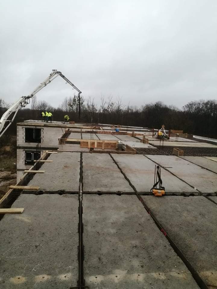 Wylewanie betonu na strop budynku w trakcie budowy z użyciem pompy do betonu; widoczni pracownicy w odblaskowych kamizelkach, szalunki i zbrojenia.