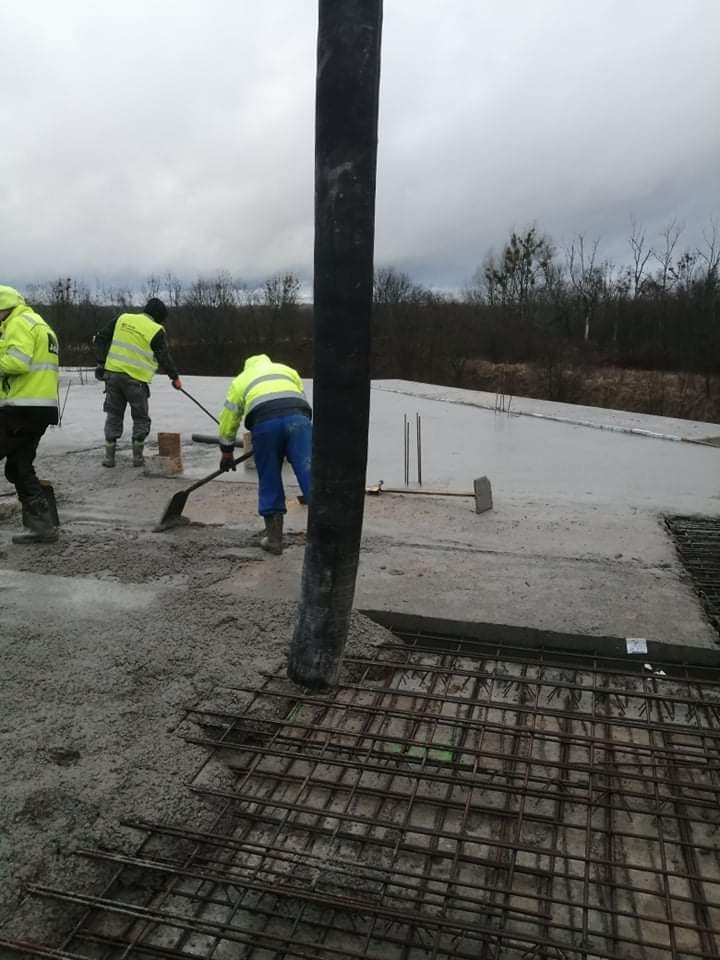 Wylewanie betonu na zbrojenie fundamentu przez pracowników w odblaskowych kamizelkach, widoczna pompa do betonu i narzędzia.