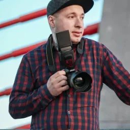 Młody mężczyzna w kraciastej koszuli i czapce z daszkiem, trzymający aparat Nikon z lampą błyskową na szyi, na tle czerwonej konstrukcji i błękitnego nieba.