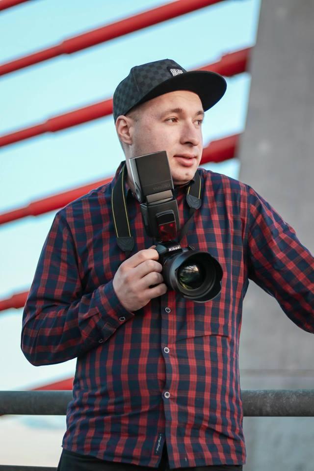 Młody mężczyzna w kraciastej koszuli i czapce z daszkiem, trzymający aparat Nikon z lampą błyskową na szyi, na tle czerwonej konstrukcji i błękitnego nieba.