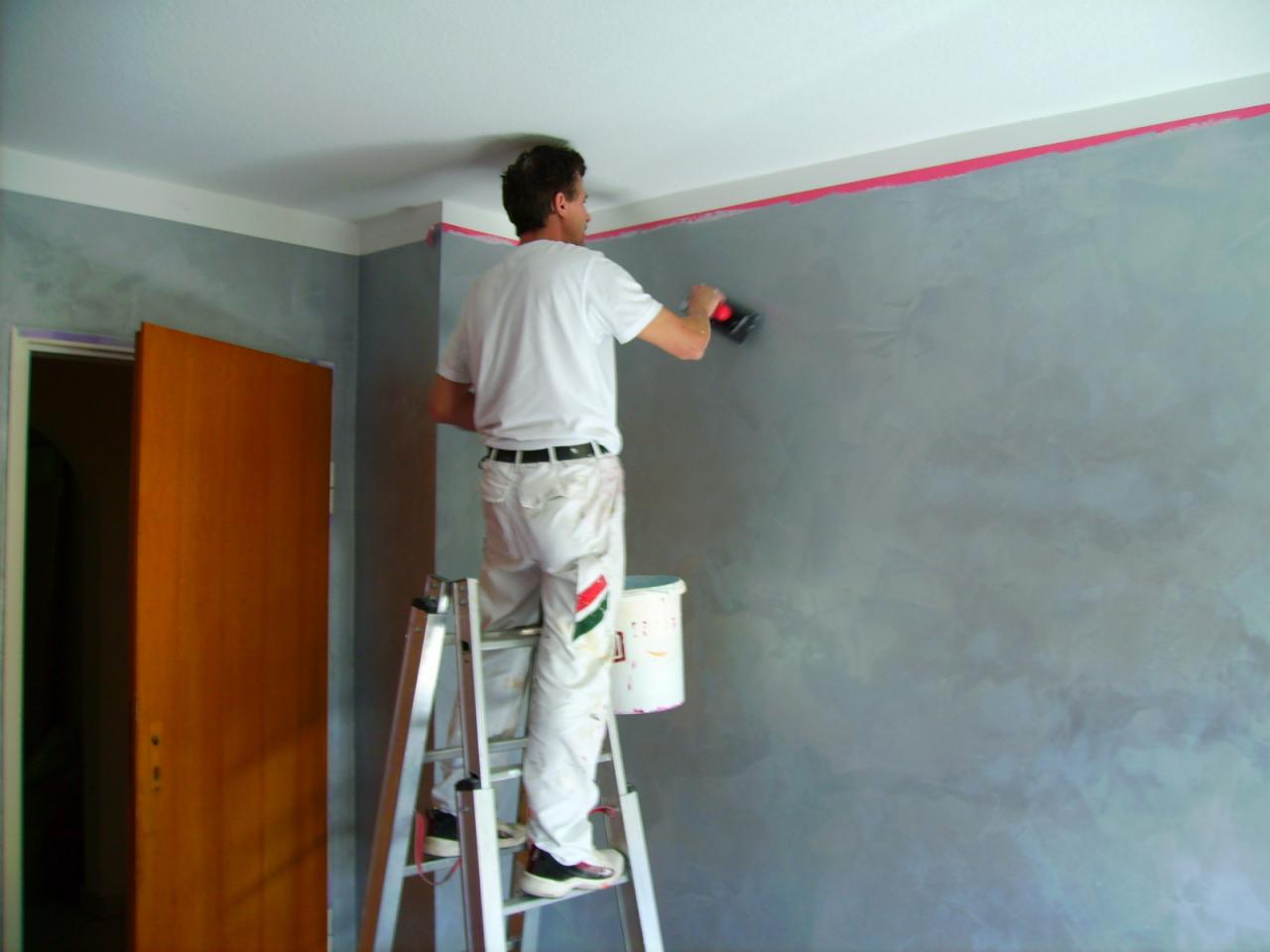 Mężczyzna na drabinie aplikuje dekoracyjną szarą farbę na ścianę za pomocą packi, zabezpieczona taśmą krawędź sufitu, widoczny otwarty drewniany futryna drzwi.