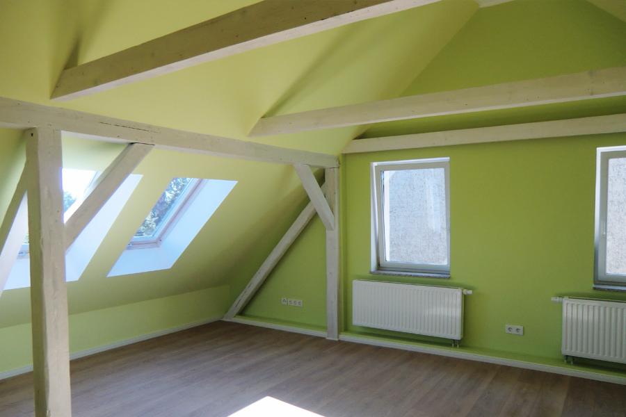 Pomalowane na jasnozielono poddasze z drewnianymi belkami pomalowanymi na biało, dwoma oknami dachowymi i dwoma zwykłymi oknami, grzejniki pod oknami, panele podłogowe