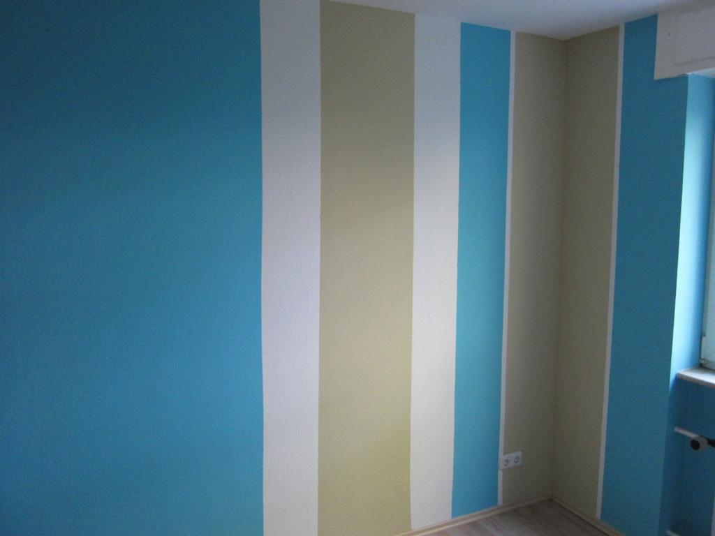 Ściana pokoju z pionowymi pasami w kolorach: błękitny, biały i beżowy, widoczny fragment okna i podłogi.