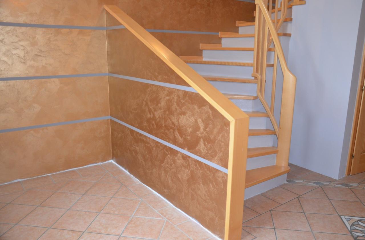 Ściana pokryta dekoracyjną, złoto-brązową farbą z poziomymi, srebrnymi pasami, przylegająca do drewnianych schodów z jasnego drewna i brązowej podłogi z płytek.