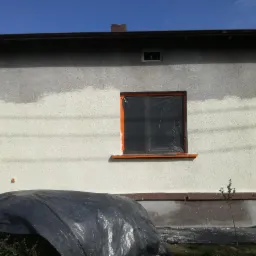 Fragment elewacji budynku z częściowo nałożoną warstwą farby, okna zabezpieczone folią, dół budynku przykryty plandeką.