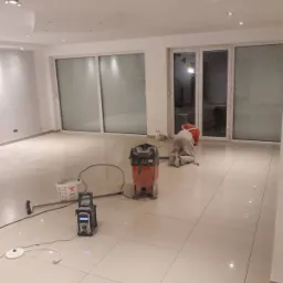 Polerowanie jasnych, błyszczących płytek podłogowych w przestronnym, jasnym pomieszczeniu z dużymi oknami przesuwnymi; widoczny pracownik w kombinezonie i sprzęt polerski.