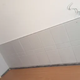 Świeżo położone, jasne, kwadratowe płytki ceramiczne na ścianie w łazience, widoczne wystające kable i niedokładne wykończenie przy suficie.