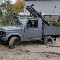 Szary samochód terenowy z wiertnicą w błotnistym terenie, widoczne ślady opon i drzewa w tle.