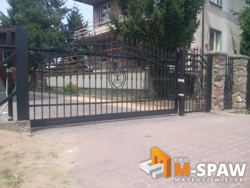 Czarna, metalowa brama przesuwna z dekoracyjnym ornamentem na tle domu jednorodzinnego i ogrodzenia z kamiennymi słupkami.
