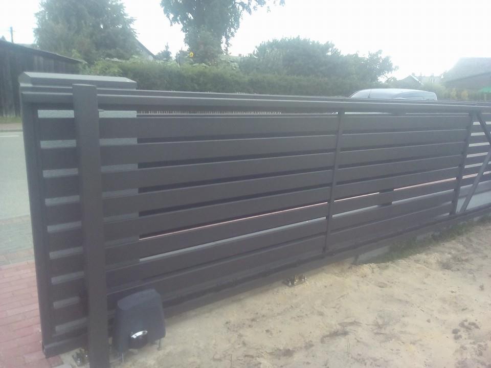 Przesuwna brama metalowa w kolorze grafitowym z automatyką, widoczna z boku, z kostką brukową i piaskiem u podstawy.