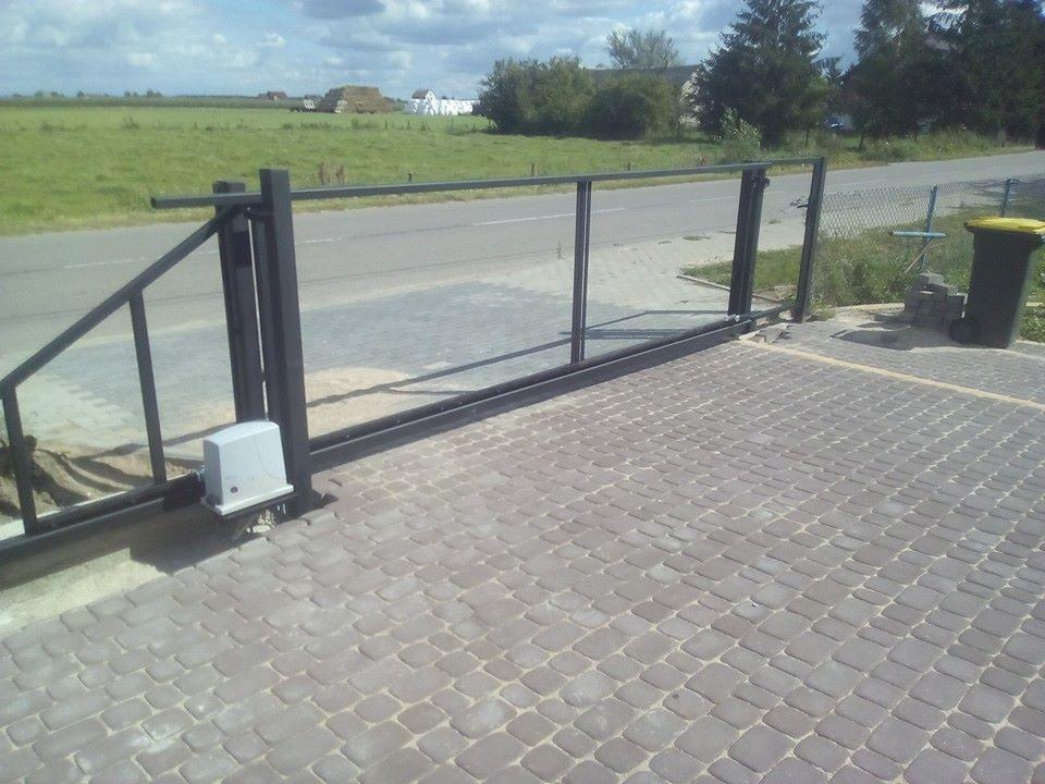 Przesuwna brama wjazdowa z automatyką, widok na szarą kostkę brukową, ulicę i pole w tle.