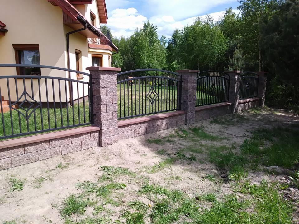 Czarne, metalowe ogrodzenie z dekoracyjnym motywem rombu, osadzone na murku z cegły klinkierowej w odcieniach różu, na tle domu i drzew.