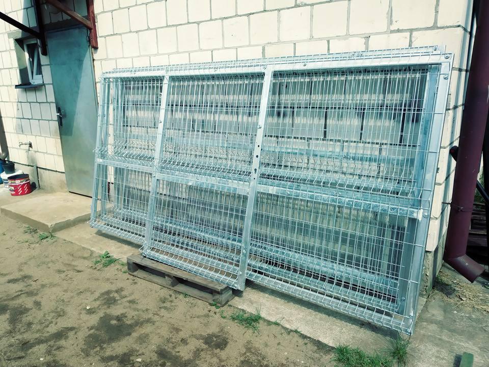 Dwa ocynkowane panele ogrodzeniowe o oczkach z drutu, oparte o ścianę budynku z białej cegły, na drewnianej palecie.