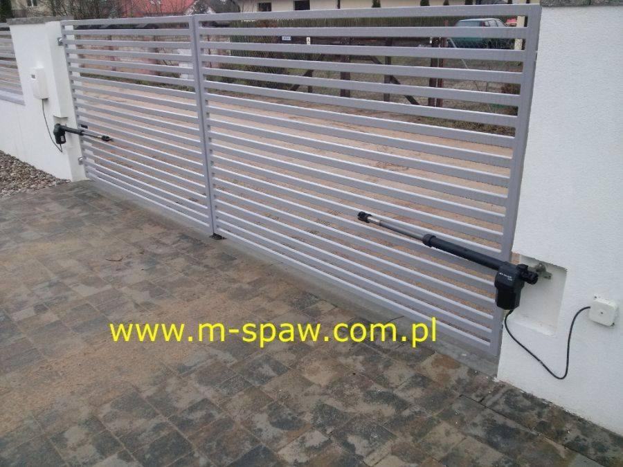 Szara brama przesuwna z automatyką, widoczne siłowniki i skrzynka sterująca na białym słupku, kostka brukowa na podjeździe.