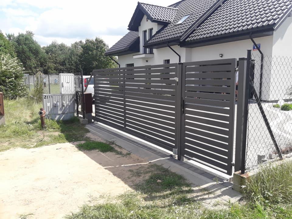 Nowoczesna, pozioma brama przesuwna i fragment ogrodzenia z metalowych profili w kolorze antracytowym na tle białego domu z ciemną dachówką.