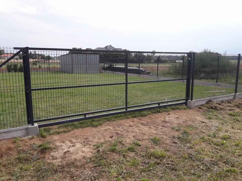 Przesuwna brama z paneli ogrodzeniowych w kolorze grafitowym, betonowa podmurówka, widok na trawnik i budynek gospodarczy w tle.