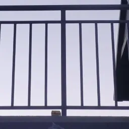 Zewnętrzna, metalowa balustrada balkonowa z pionowymi prętami i poziomymi poręczami, pomalowana na ciemny kolor, z widokiem na jasne niebo i fragment budynku w tle.