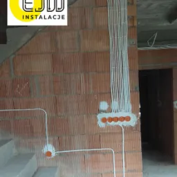 Surowe wnętrze w trakcie prac: widoczne kable elektryczne w peszlach, puszki instalacyjne w ścianie z cegły dziurawki, logo firmy instalacyjnej w lewym górnym rogu.