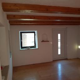 Robert Dawiec - Wnętrze pokoju po remoncie: drewniane belki stropowe, okno z ozdobną szybą, białe ściany z wnękami, drewniana podłoga i białe drzwi.