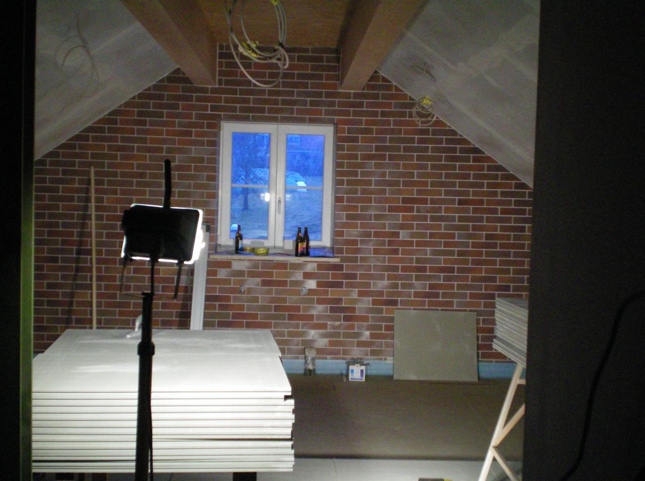 Poddasze z oknem, ceglaną ścianą i stosem płyt kartonowo-gipsowych, oświetlone lampą budowlaną, z butelkami na parapecie.