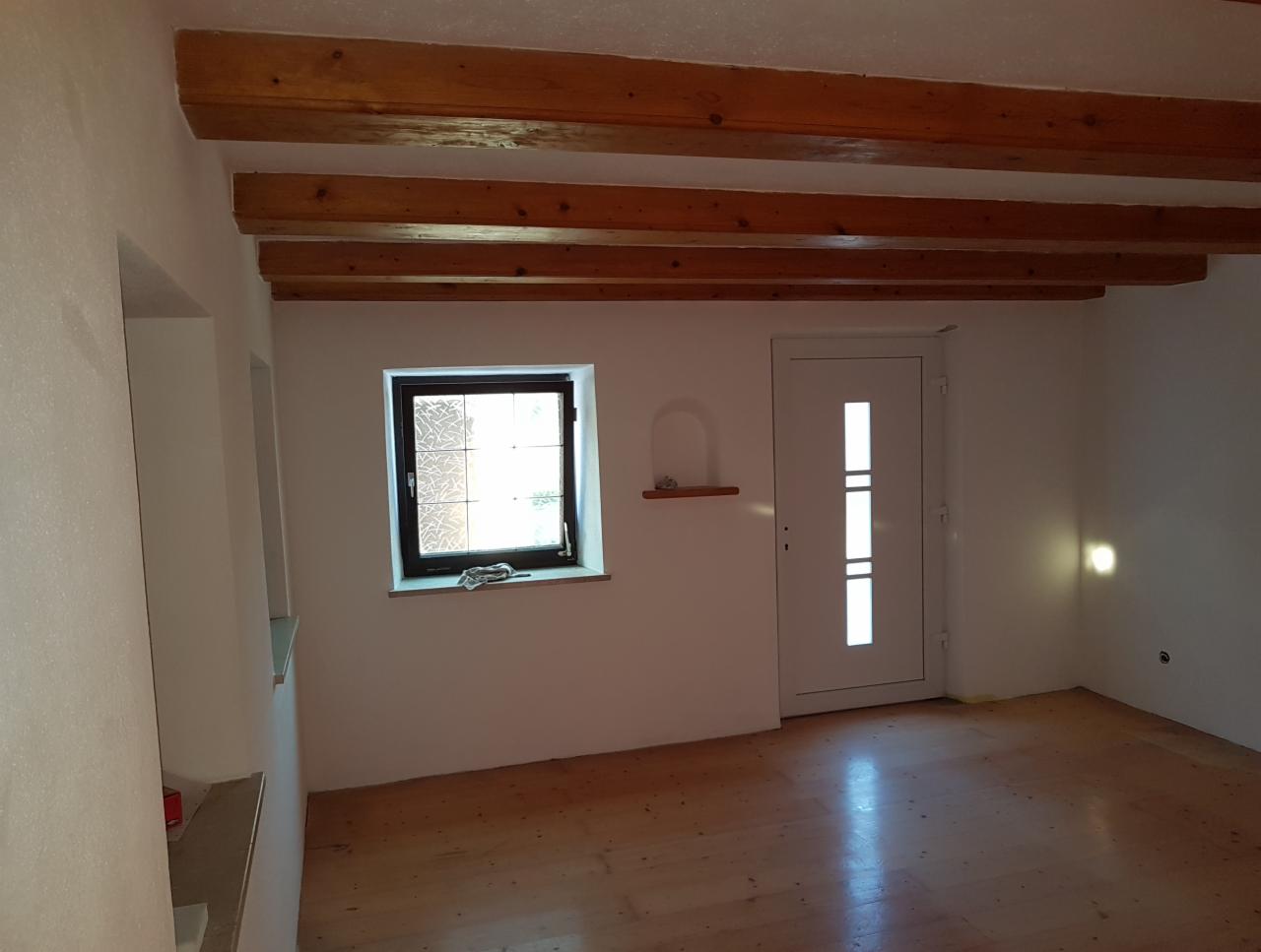Wnętrze pokoju po remoncie: drewniane belki stropowe, okno z ozdobną szybą, białe ściany z wnękami, drewniana podłoga i białe drzwi.