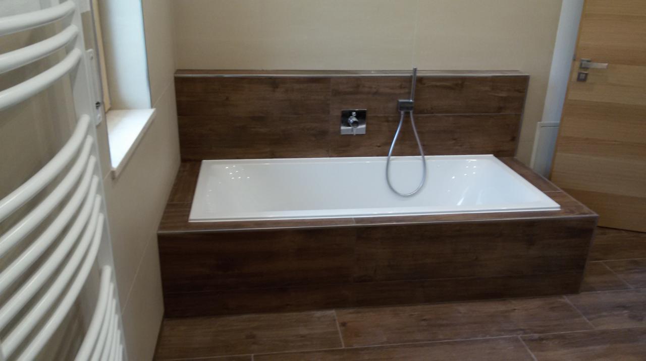 Nowoczesna łazienka z białą wanną obudowaną panelami imitującymi ciemne drewno, dopasowanymi do płytek podłogowych, oraz białym grzejnikiem drabinkowym.