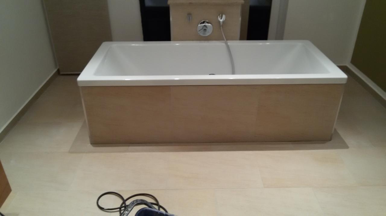 Wanna obudowana płytkami imitującymi drewno, armatura zamontowana w ścianie, jasna podłoga z płytek, fragment narzędzia w rogu kadru.