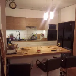 Nowoczesna kuchnia z białymi i drewnianymi frontami, wyspą z hokerami, oświetleniem punktowym i czarną lodówką, widok na blat z podkładkami, szklanką i kieliszkiem wina.