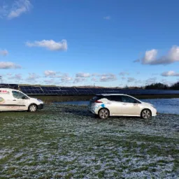 Dwa samochody elektryczne firmy Rawicom.pl stoją na trawie z panelami słonecznymi w tle, widoczne odbicie paneli w wodzie.