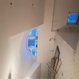 Wnętrze w trakcie remontu: białe ściany i sufit po gładziowaniu, widoczne okno półokrągłe i małe kwadratowe, drabina, resztki materiałów budowlanych na podłodze, przygotowania do dalszych prac...