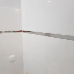 Białe, błyszczące płytki ścienne z metalową listwą dekoracyjną na wysokości wzroku, widoczny włącznik światła.