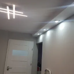 Korytarz po remoncie z nowoczesnym oświetleniem sufitowym: lampa krzyżowa i rząd halogenów w listwie, widoczna biała ściana, białe drzwi z poziomymi przeszkleniami i fragment regału w głębi.