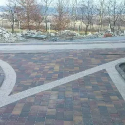 Wzór z kostki brukowej w odcieniach brązu i szarości, z elementami dekoracyjnymi z jaśniejszego kruszywa, obrzeża z kostki granitowej, w tle hałda kamieni i drzewa bez liści.