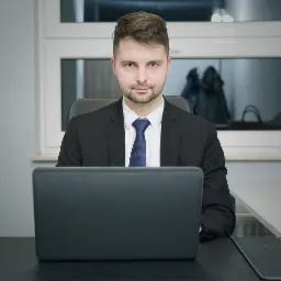 Młody mężczyzna w garniturze i krawacie siedzi za biurkiem z laptopem, spoglądając wprost w obiektyw. W tle okno i elementy wystroju biura.