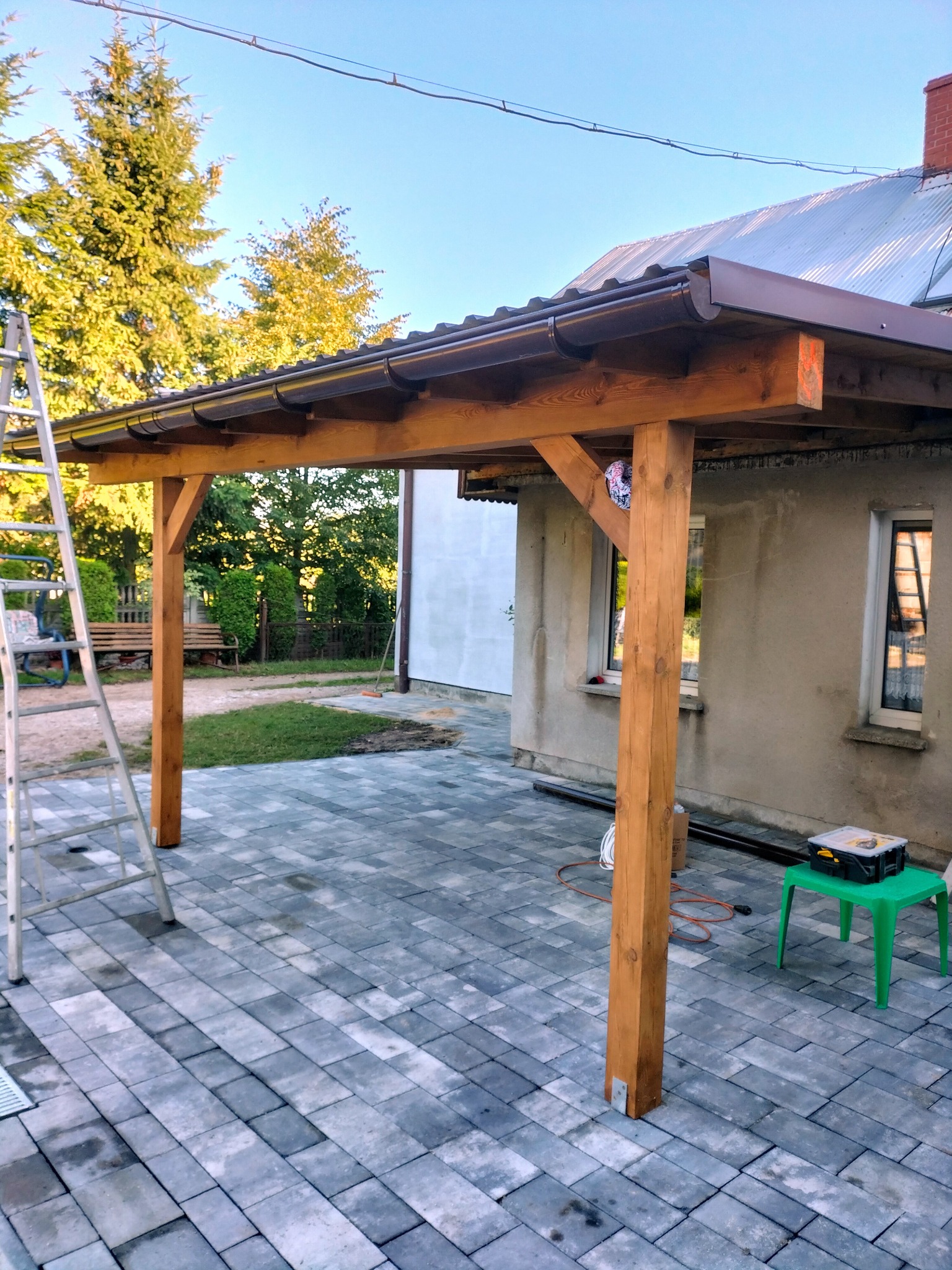 Drewniana konstrukcja z brązowym dachem, prawdopodobnie wiata lub pergola, w trakcie budowy, z widoczną drabiną, narzędziami i kostką brukową na podłożu.