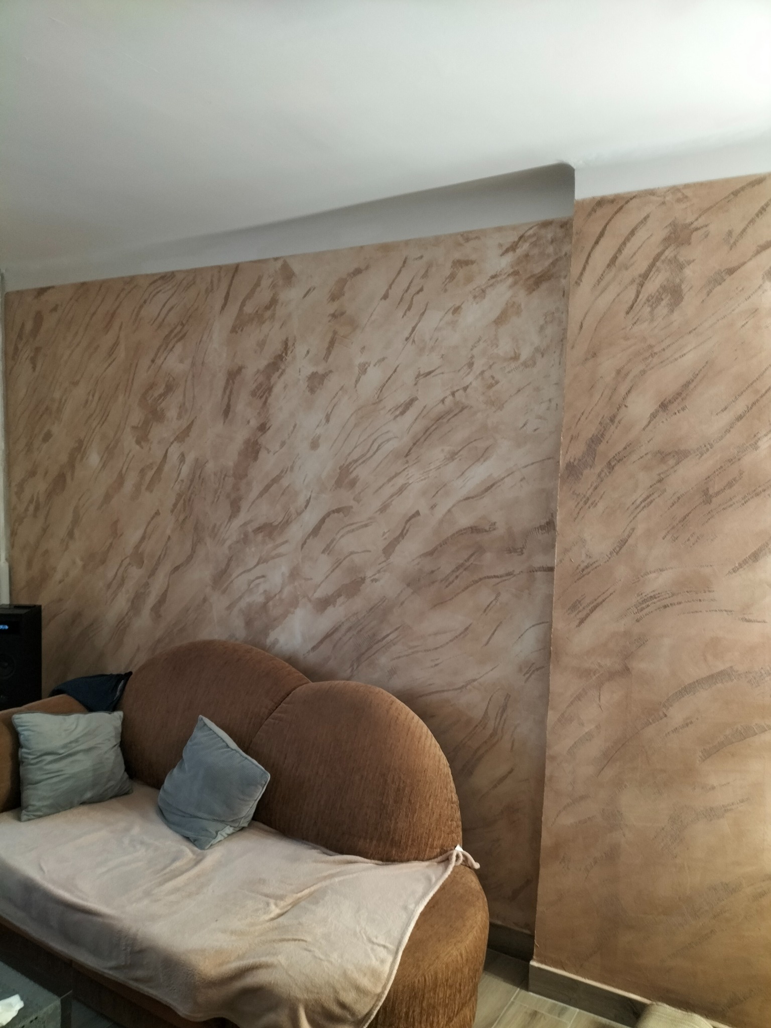 Ściana w salonie z dekoracyjną strukturą w odcieniach beżu i brązu, widoczny fragment sofy z poduszkami.