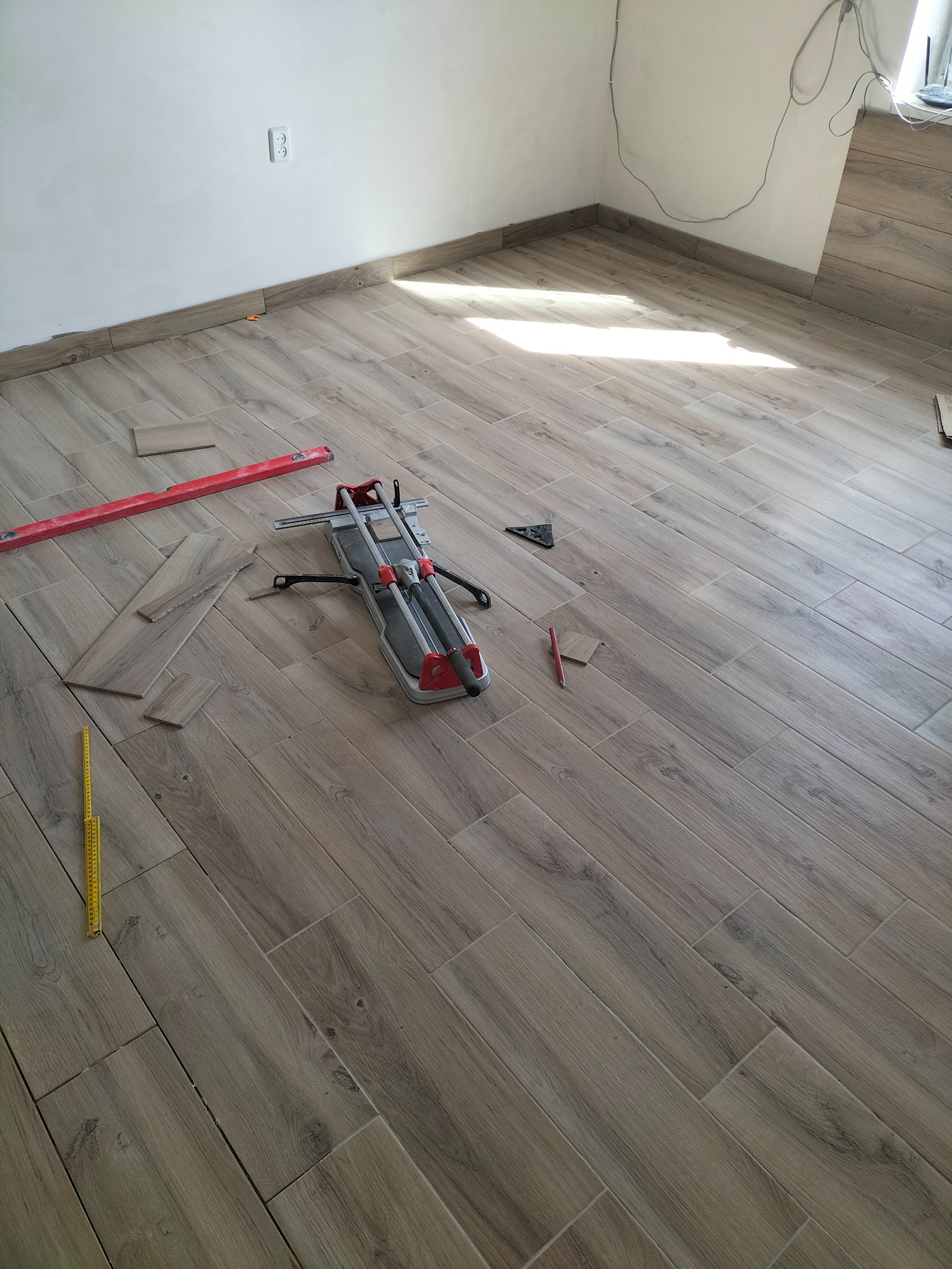 Ułożona pod kątem podłoga z płytek imitujących drewno, narzędzia glazurnicze: przecinarka do płytek, poziomica, kątownik, miarka i ołówek.